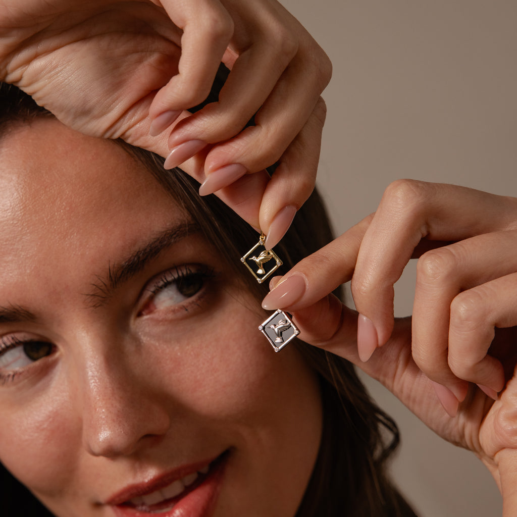 A woman smiles softly, holding two Onyx Hummingbird Charms with engraved letters close to her face.