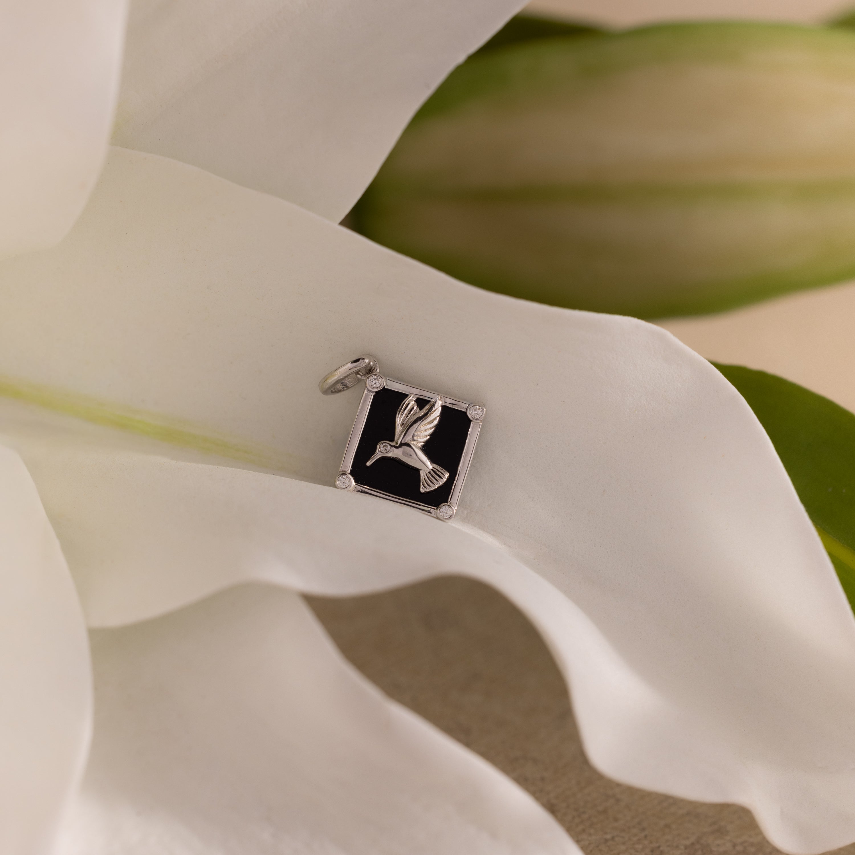 The Onyx Hummingbird Charm in Silver rests on a black square atop soft white flower petals and green leaves—a refined piece to elevate your jewelry collection.