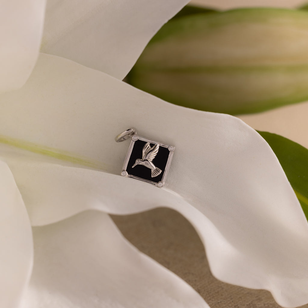 The Onyx Hummingbird Charm, a silver bird pendant, rests on a white flower with green leaves in the background.