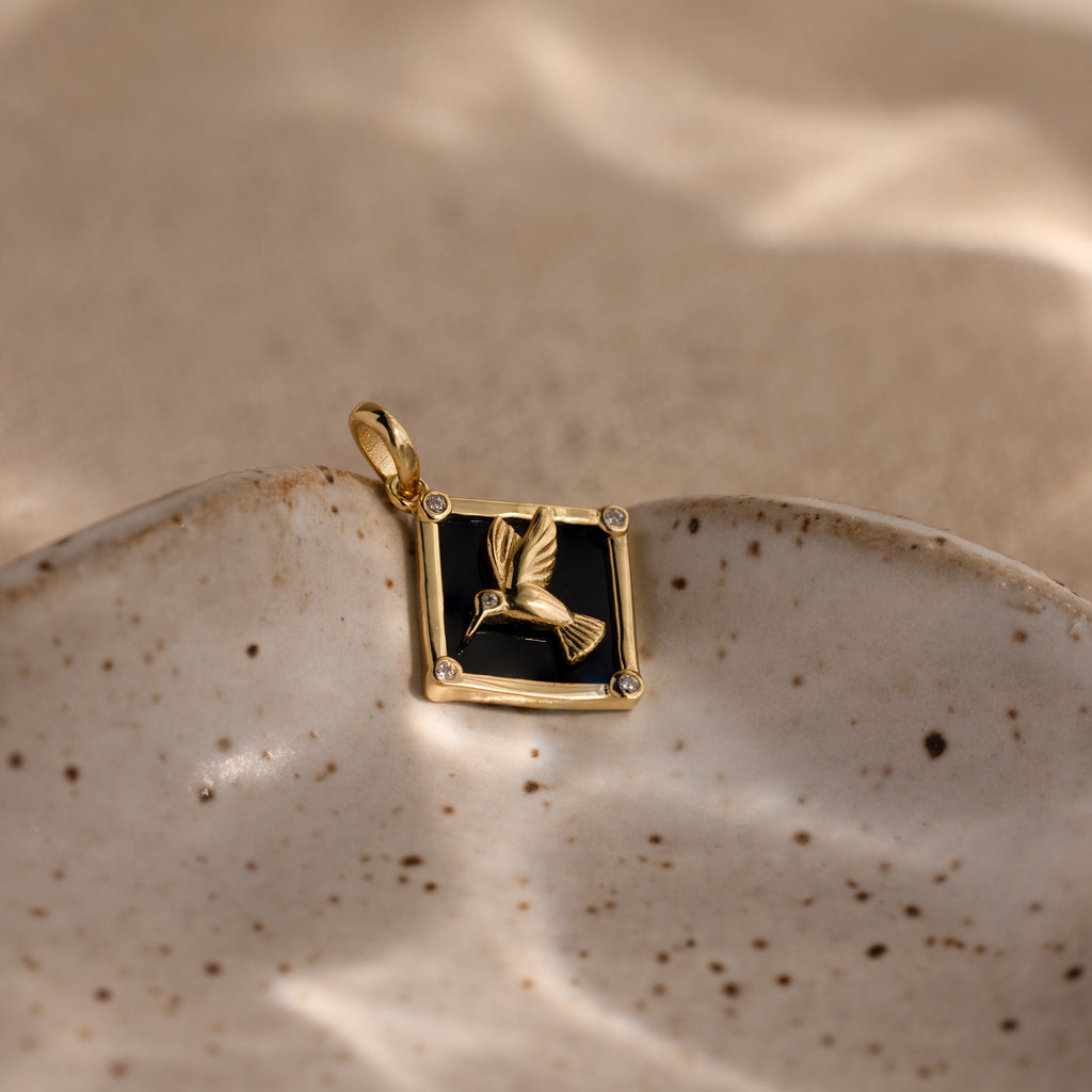 The Onyx Hummingbird Charm in 18K Gold glows against a black background, elegantly presented on a speckled ceramic dish under soft light.