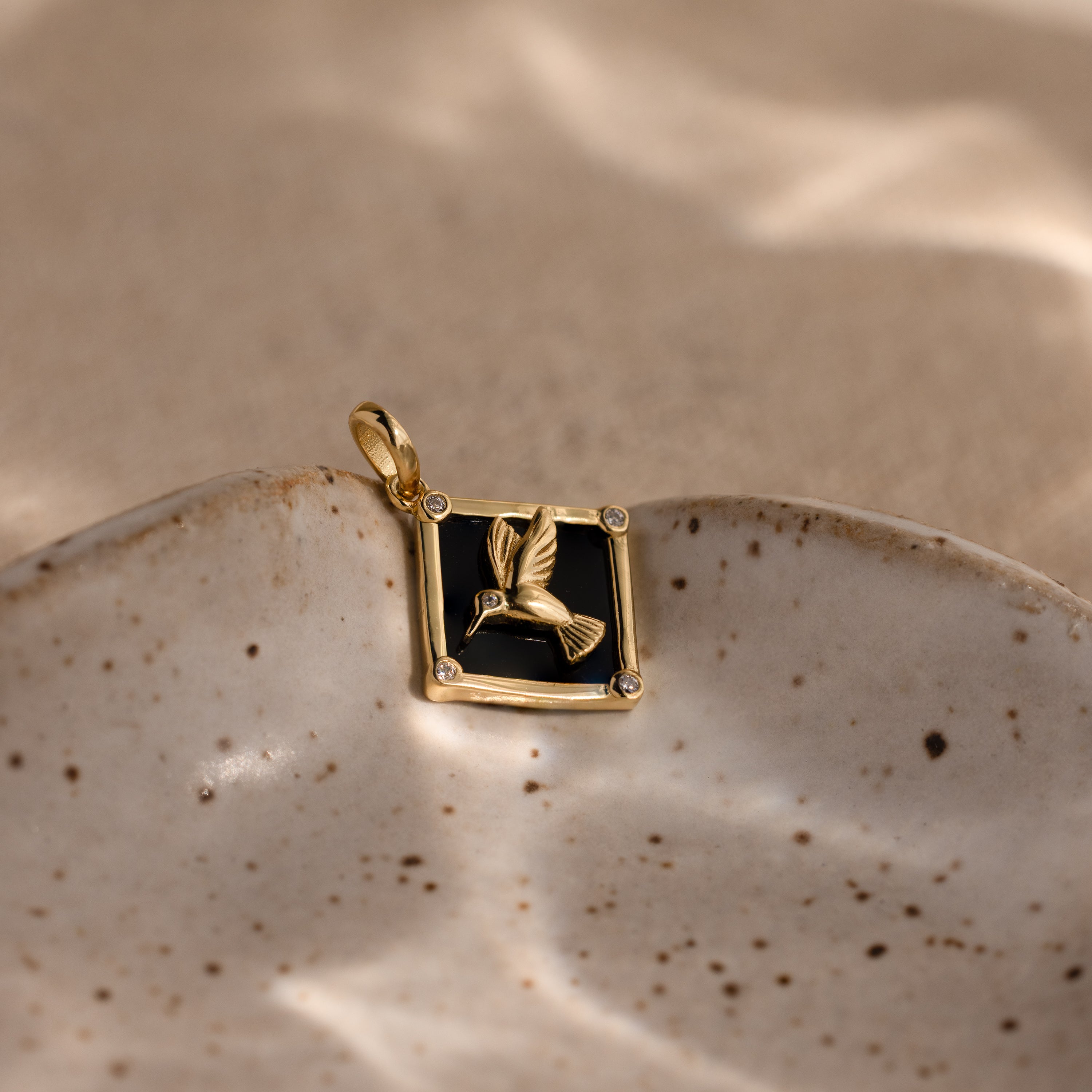 The Onyx Hummingbird Charm in 18K Gold glows against a black background, elegantly presented on a speckled ceramic dish under soft light.