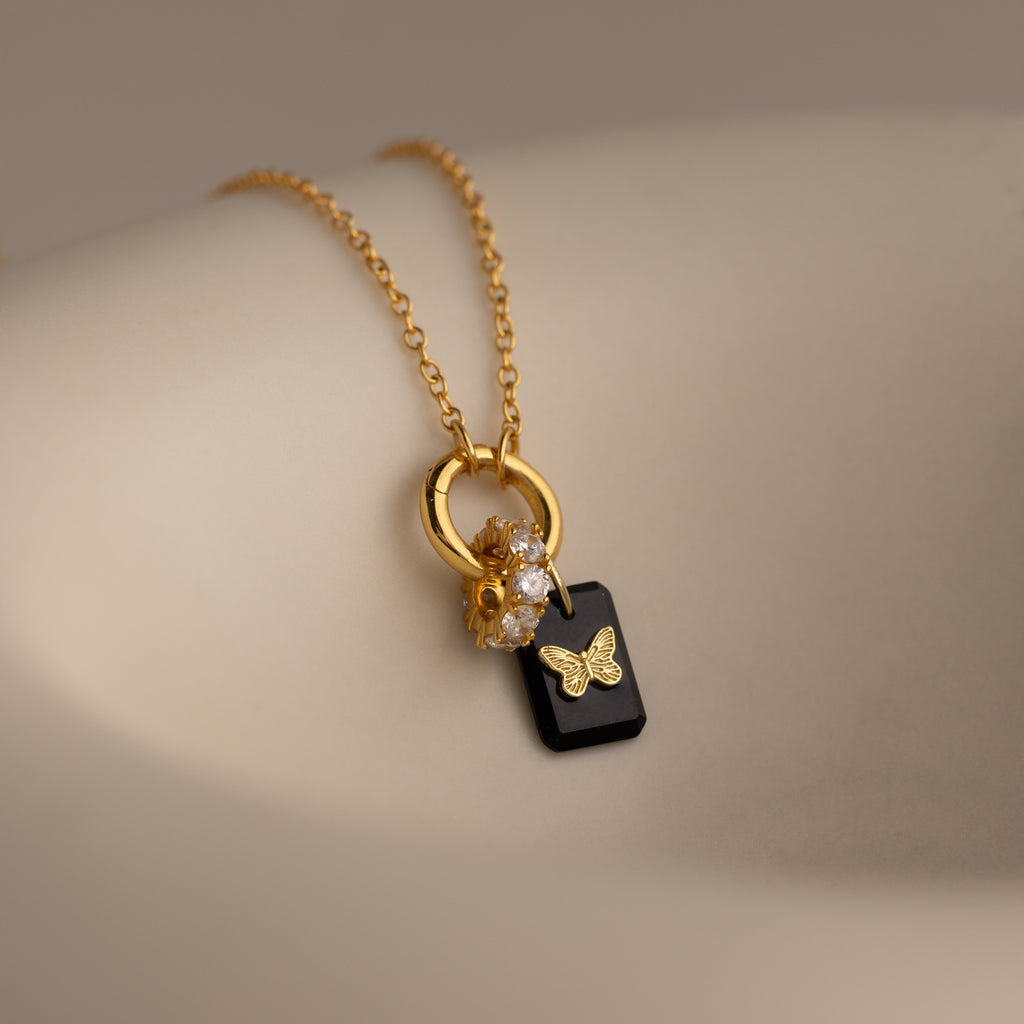 Onyx Butterfly Charm gold necklace featuring a delicate butterfly pendant and sparkling crystal charms, elegantly displayed on a soft beige surface.