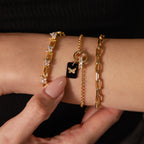 A close-up of a wrist adorned with three gold bracelets, including the Onyx Butterfly Charm in 18K Gold, as a hand gently touches the wrist—radiating elegance and charm.