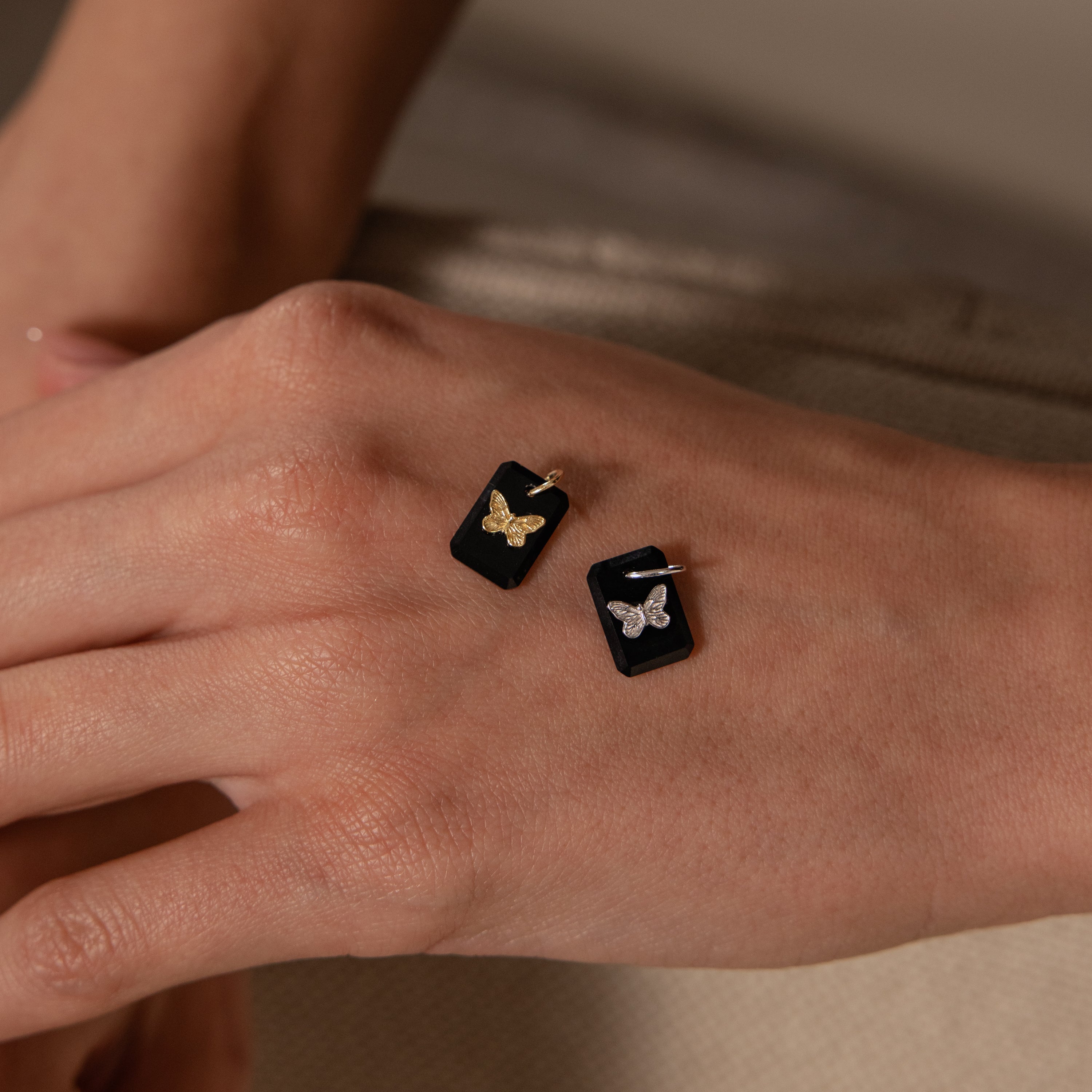 A hand wears two black rectangular Onyx Butterfly Charm rings, each featuring a butterfly ornament—one in gold, one in silver.