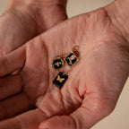 A hand holds three Onyx Butterfly Charms in 18K Gold—small gold and black pendants, each intricately designed.