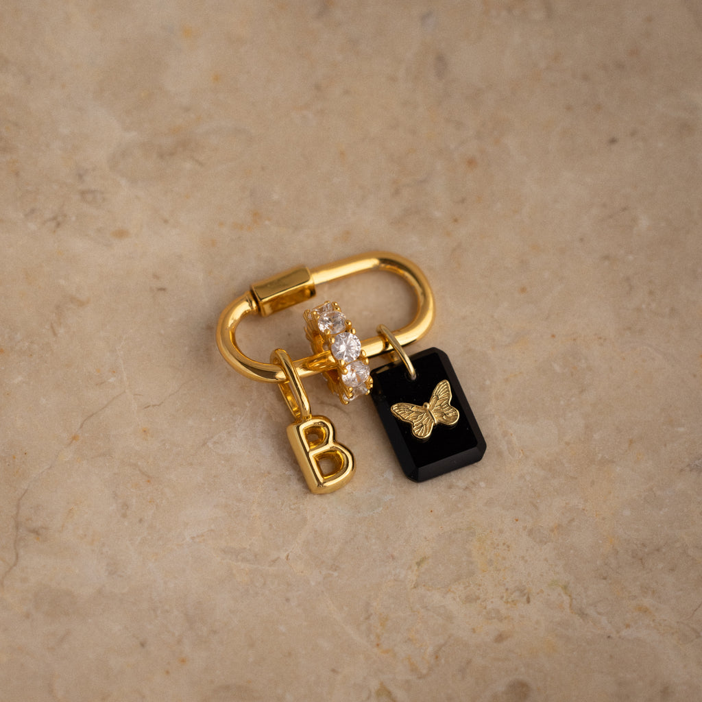 The Onyx Butterfly Charm in 18K Gold features a “B” charm, clear gem ring, and a black tag with a gold butterfly, adding personalized elegance to your keys or bag.