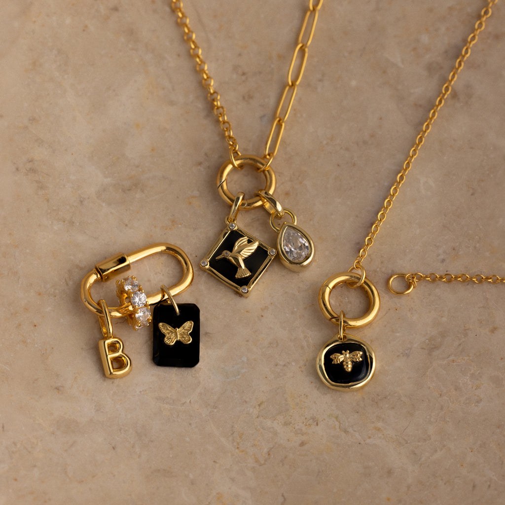 Three gold necklaces feature various charms such as letters, butterflies, birds, and a teardrop-shaped gem—including the Onyx Hummingbird Charm.