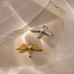 Two Angel Wing Charms—one gold, one silver—each with a heart-shaped center, rest on a feathered surface. These pendants are ideal as protection jewelry or a meaningful gift for someone special.