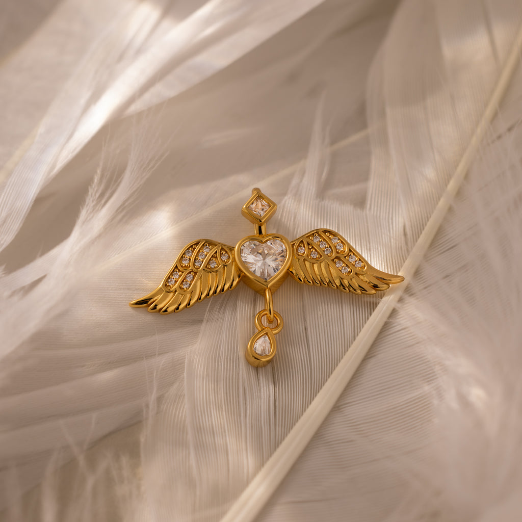 The Angel Wing Charm in 18K Gold features pavé-set wings and a heart-shaped gem, elegantly displayed on soft white feathers.