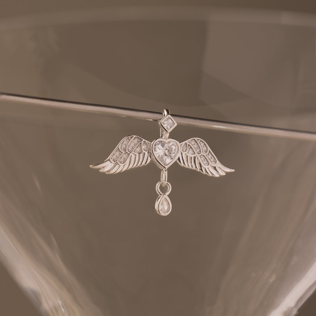 The Angel Wing Charm in Silver features pavé-set wings and a crystal drop, displayed hanging on the rim of a clear glass.