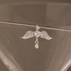 The Angel Wing Charm in Silver features pavé-set wings and a crystal drop, displayed hanging on the rim of a clear glass.