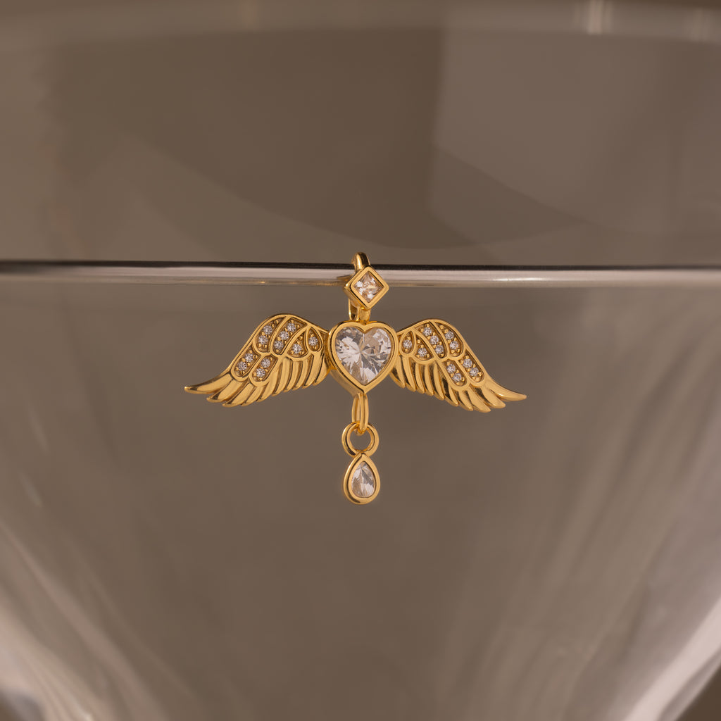 The Angel Wing Charm in 18K Gold, with pavé-set wings and diamond accents plus a drop diamond, hangs elegantly on the edge of a transparent glass surface.