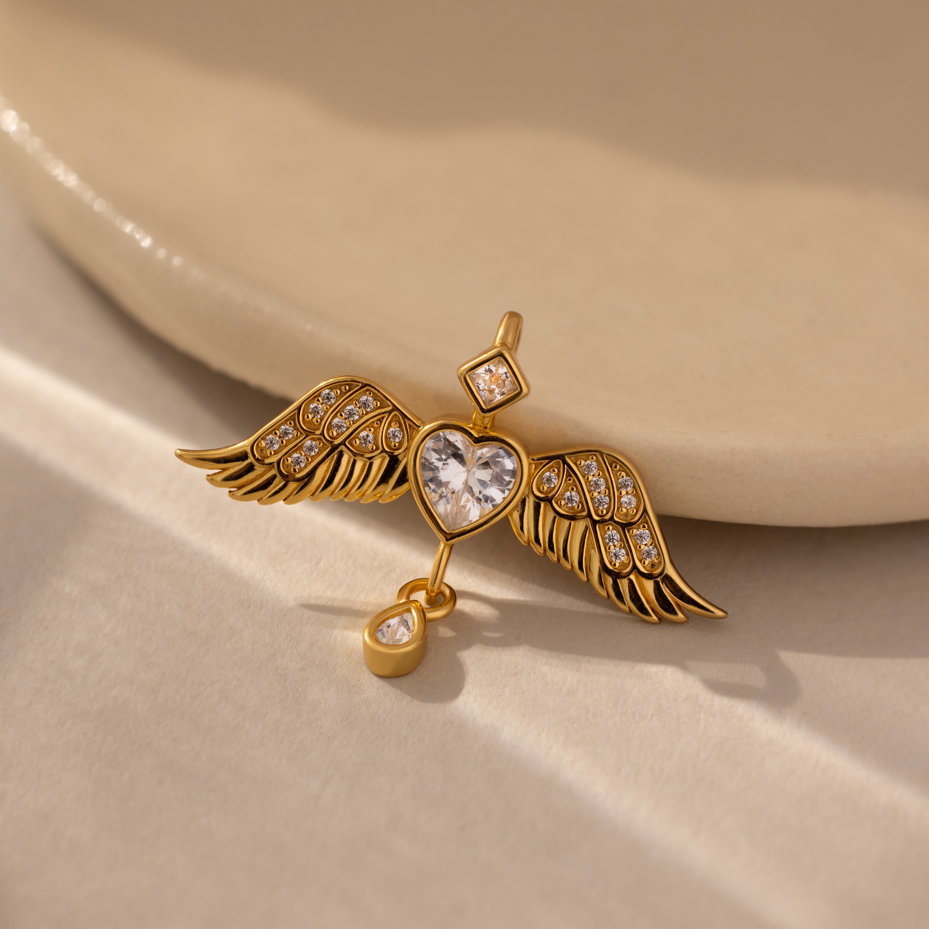 Angel Wing Charm in 18K Gold features pavé-set wings, a heart-shaped crystal center, and a small drop diamond, shown on a neutral background.
