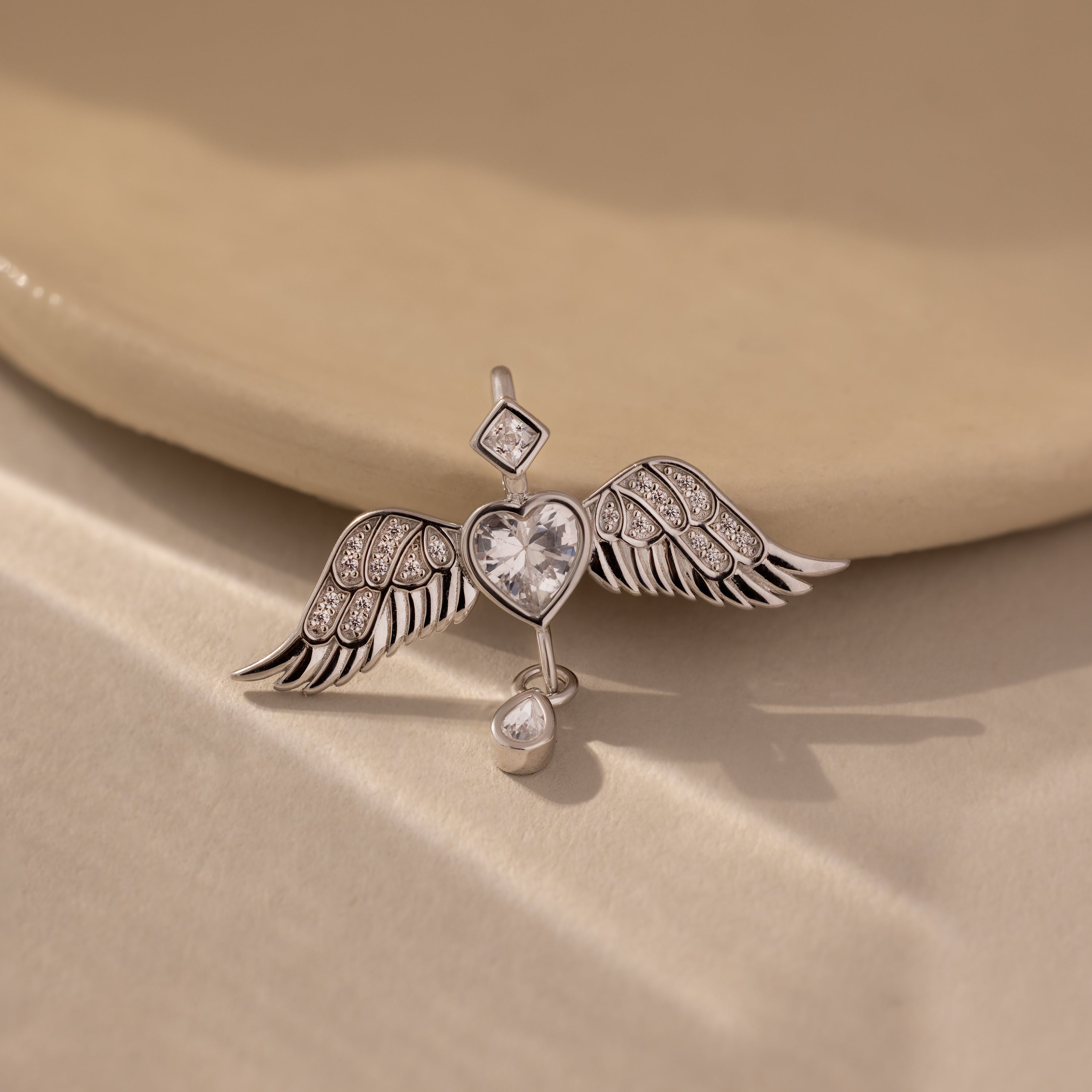 The Angel Wing Charm in Silver, featuring pavé-set crystal wings, is displayed on a beige ceramic surface bathed in soft light.
