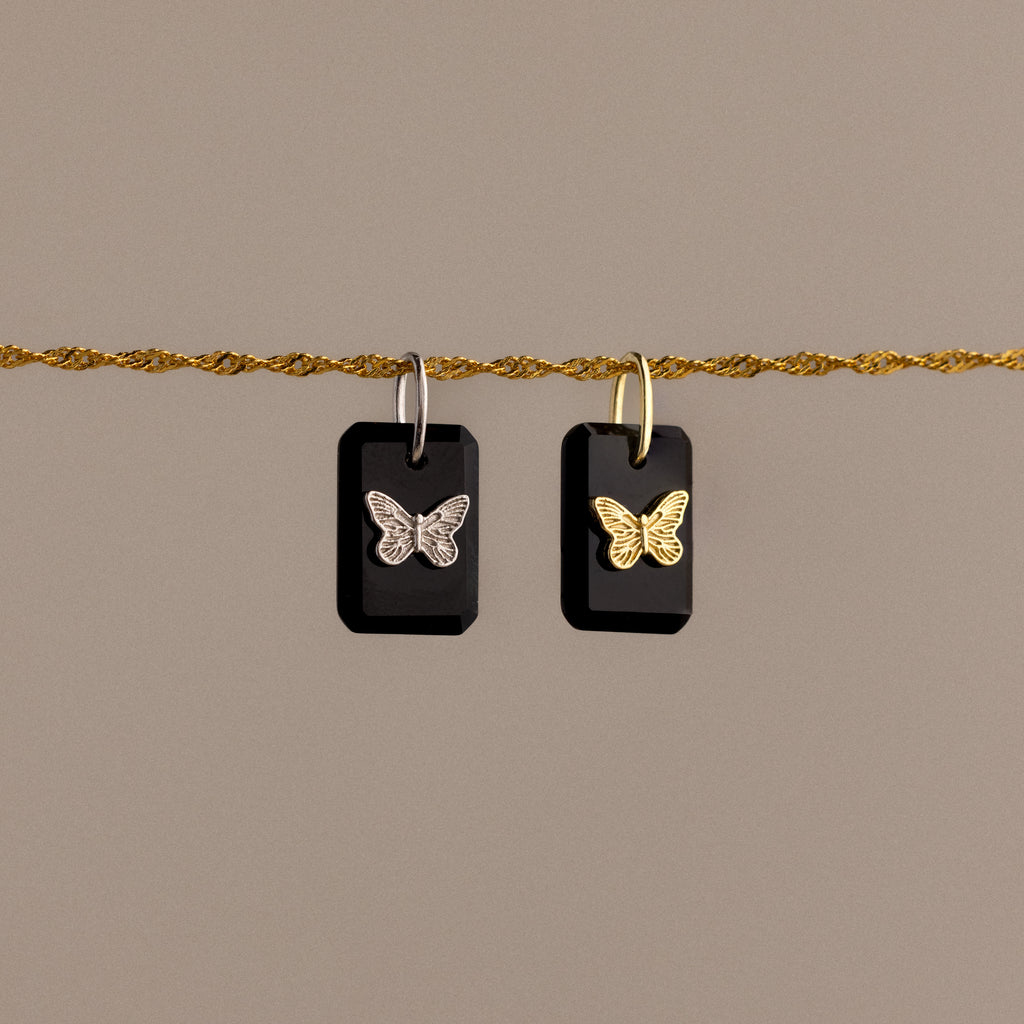 Two black rectangular Onyx Butterfly Charms with butterfly designs hang from a twisted gold rope against a beige background.