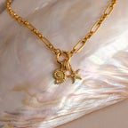 The Rolo Circle Figaro Necklace features daisy and starfish charms, displayed on a shimmering shell background for an elegant update to the classic link necklace.