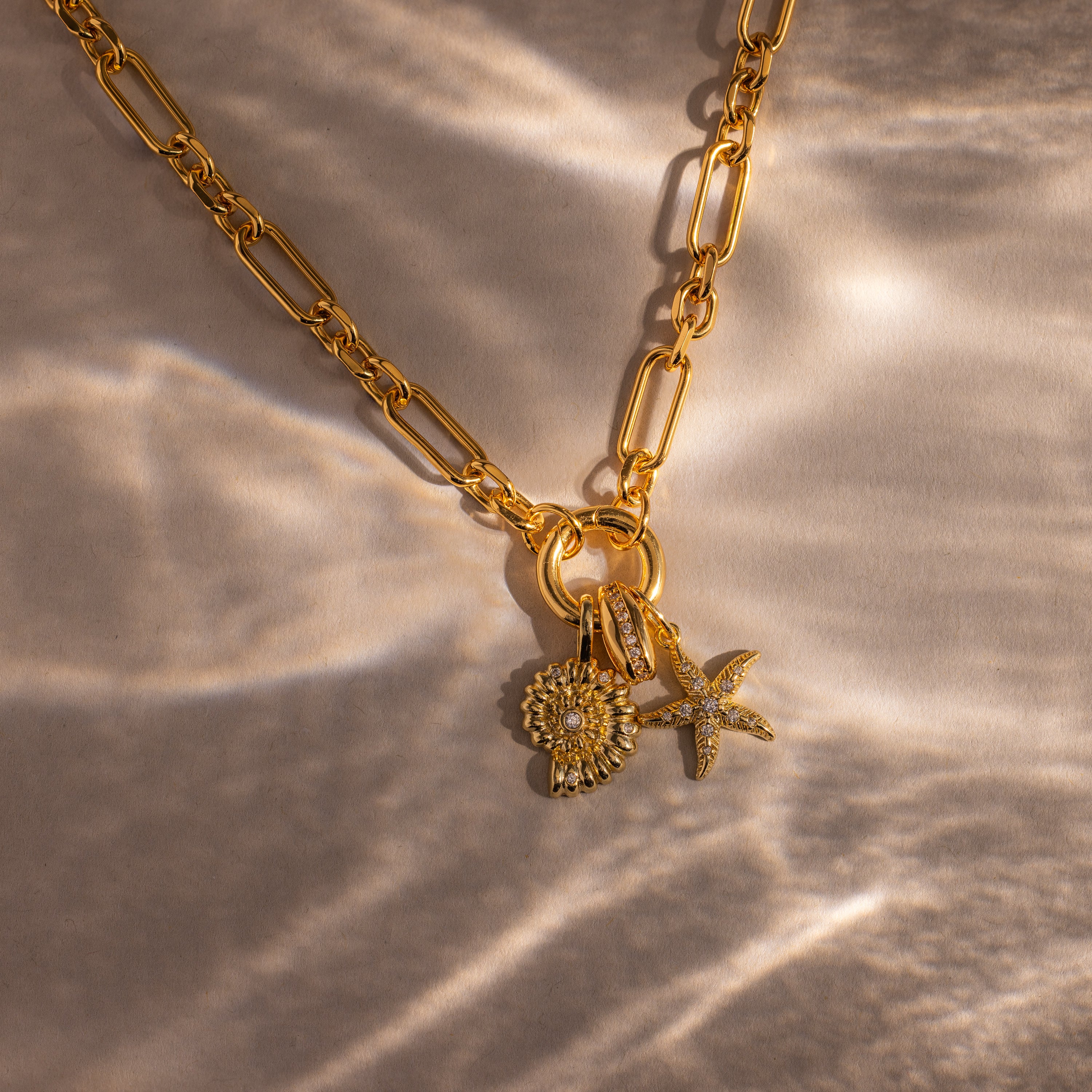 The Pave Dome Charm gold chain necklace features a stack of sun, shell, and starfish charms on a textured beige surface—an ideal accessory for fans of delicate jewelry.
