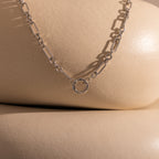 The Rolo Circle Figaro Necklace in Silver, featuring an open circle pendant and shown on smooth beige surfaces, creates a striking statement piece.