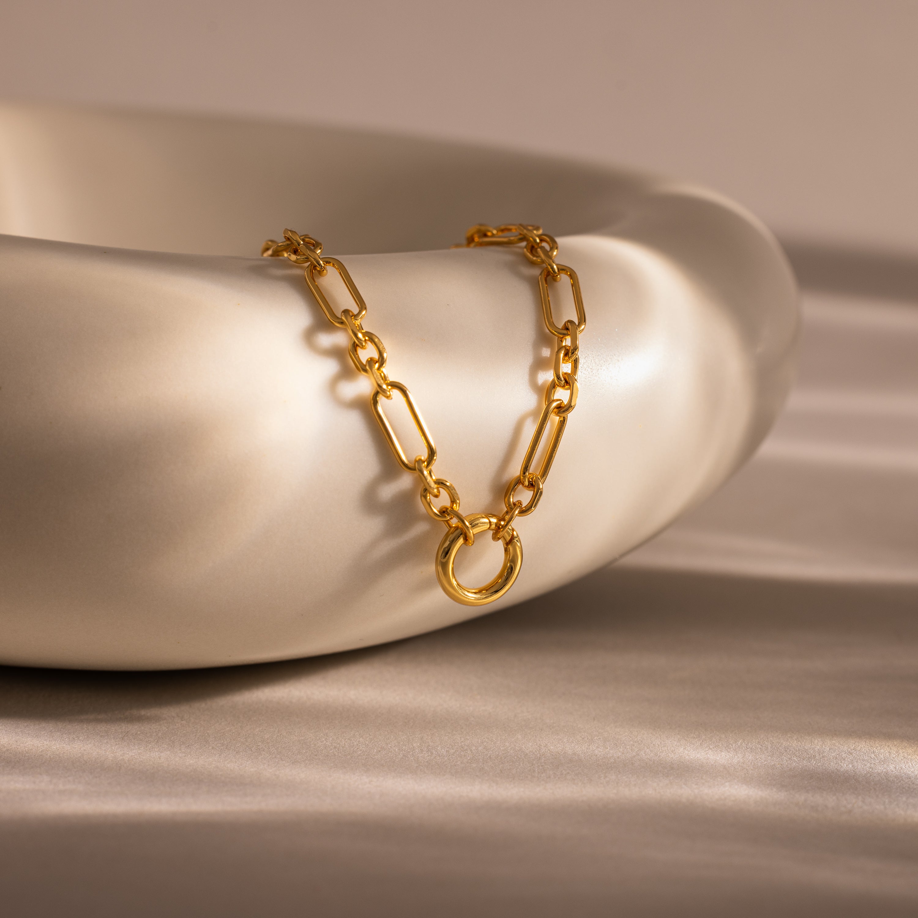 The Rolo Circle Figaro Necklace in 18K Gold is draped over a smooth, white ceramic bowl on a silky fabric surface.
