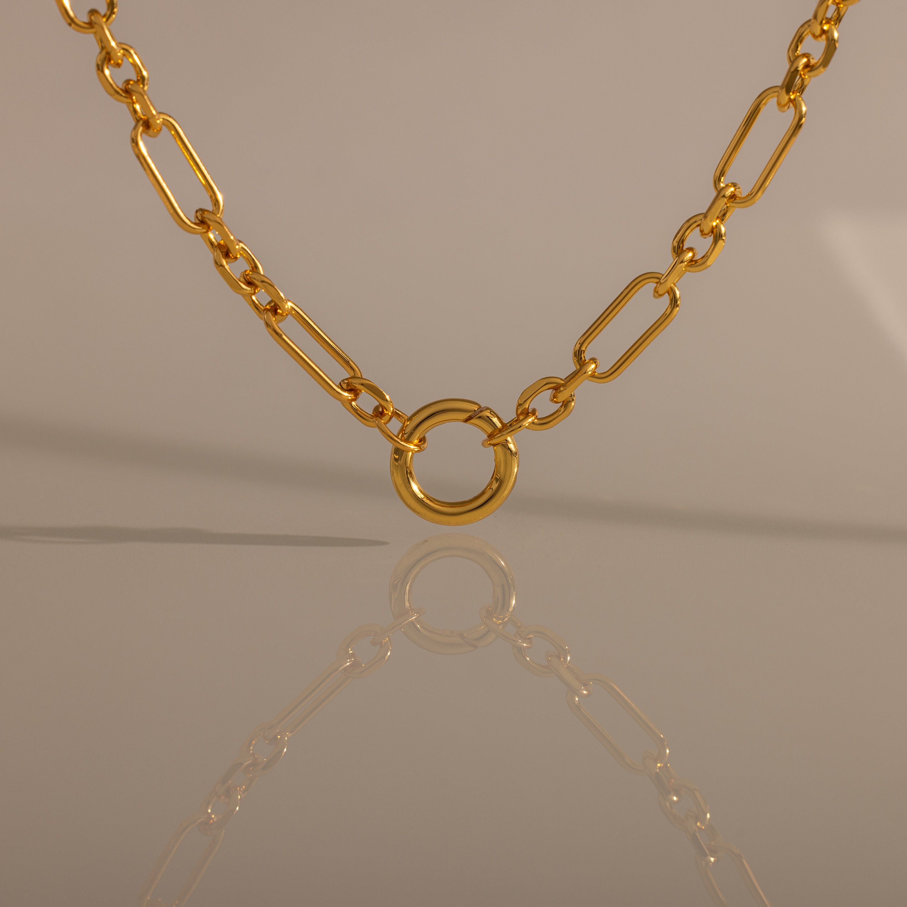 The Rolo Circle Figaro Necklace, a gold chain with a circular pendant, is reflected on a smooth beige surface for a look of effortless elegance.