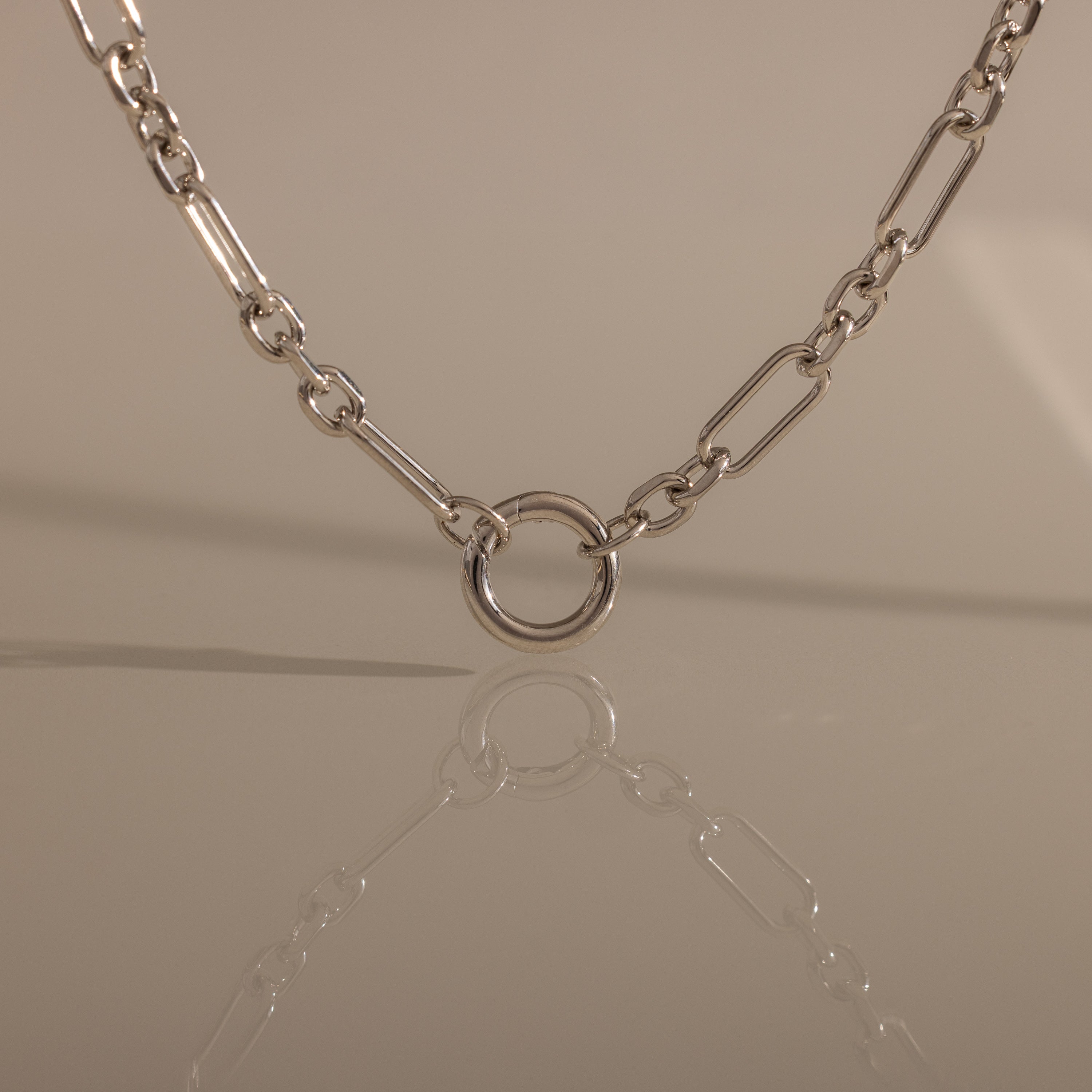 The Rolo Circle Figaro Necklace in Silver features bold links and a circular pendant, beautifully reflected on a smooth surface—a true statement piece for any jewelry collection.