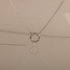 The Cable Circle Charm Necklace in Silver features a simple silver chain with a small circular pendant, elegantly reflected on a smooth surface.