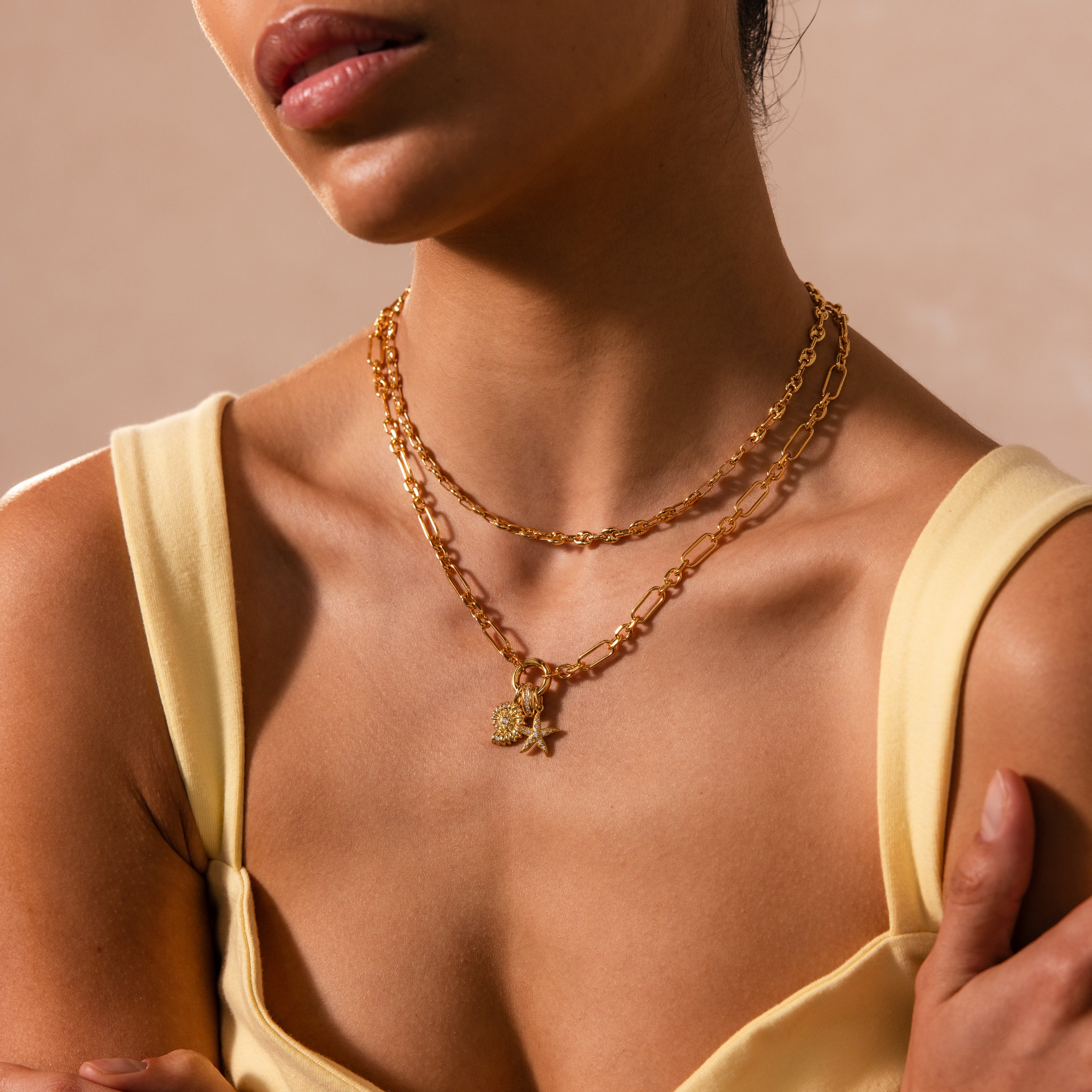 A woman wears layered 18K gold necklaces, featuring the Pavé Dome Charm in 18K Gold, which adds subtle sparkle to her look. She is dressed in a pale yellow top, shown from shoulders to lips against a neutral background.