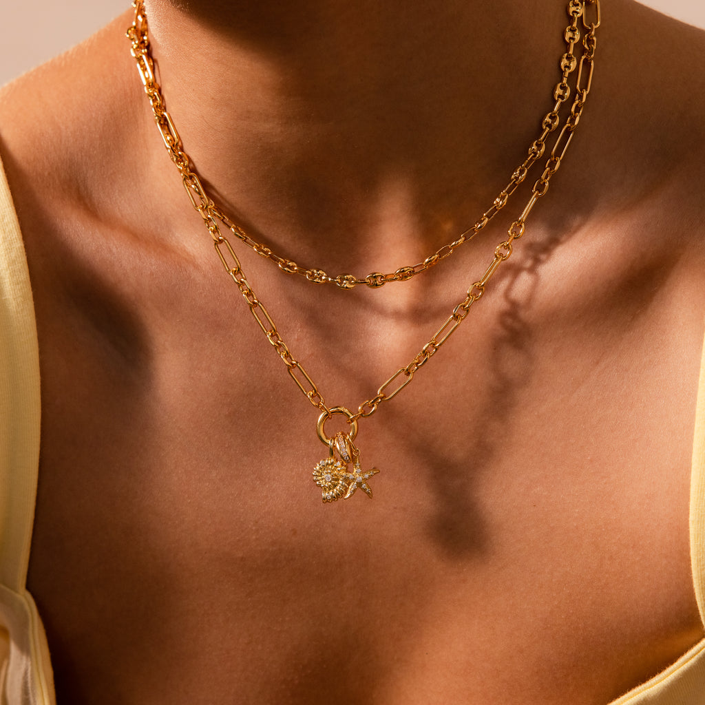 A person wearing a stack of gold chain necklaces featuring shell, starfish, and the Pave Dome Charm in Silver pendants, paired with a yellow top for extra sparkle.