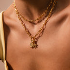 Close-up of a person wearing the Rolo Circle Figaro Necklace, a layered gold chain featuring a statement front clasp and classic links, adorned with star and shell charms on bare skin.