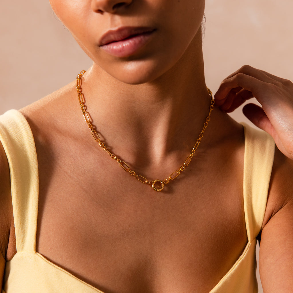Woman in a yellow top wears the Rolo Circle Figaro Necklace in 18K Gold, featuring a statement front clasp and oval links, with her hand near her neck against a neutral background.