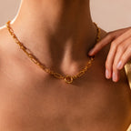 A person wearing the Rolo Circle Figaro Necklace, a gold chain statement piece with a circular design, touches their neck with one hand.