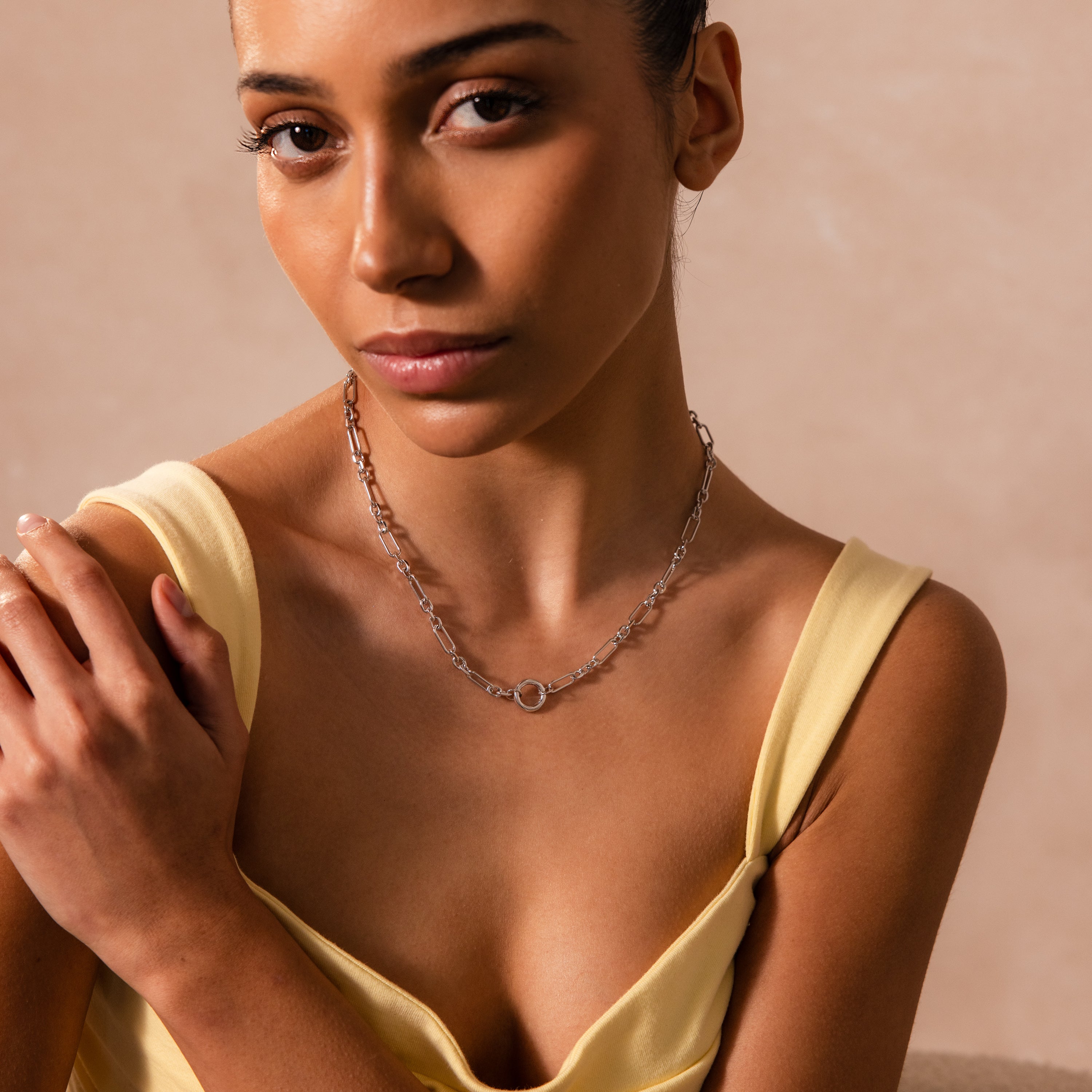 A woman in a yellow dress wears the Rolo Circle Figaro Necklace in Silver, its distinctive links adding subtle elegance as she gazes at the camera with a neutral expression.