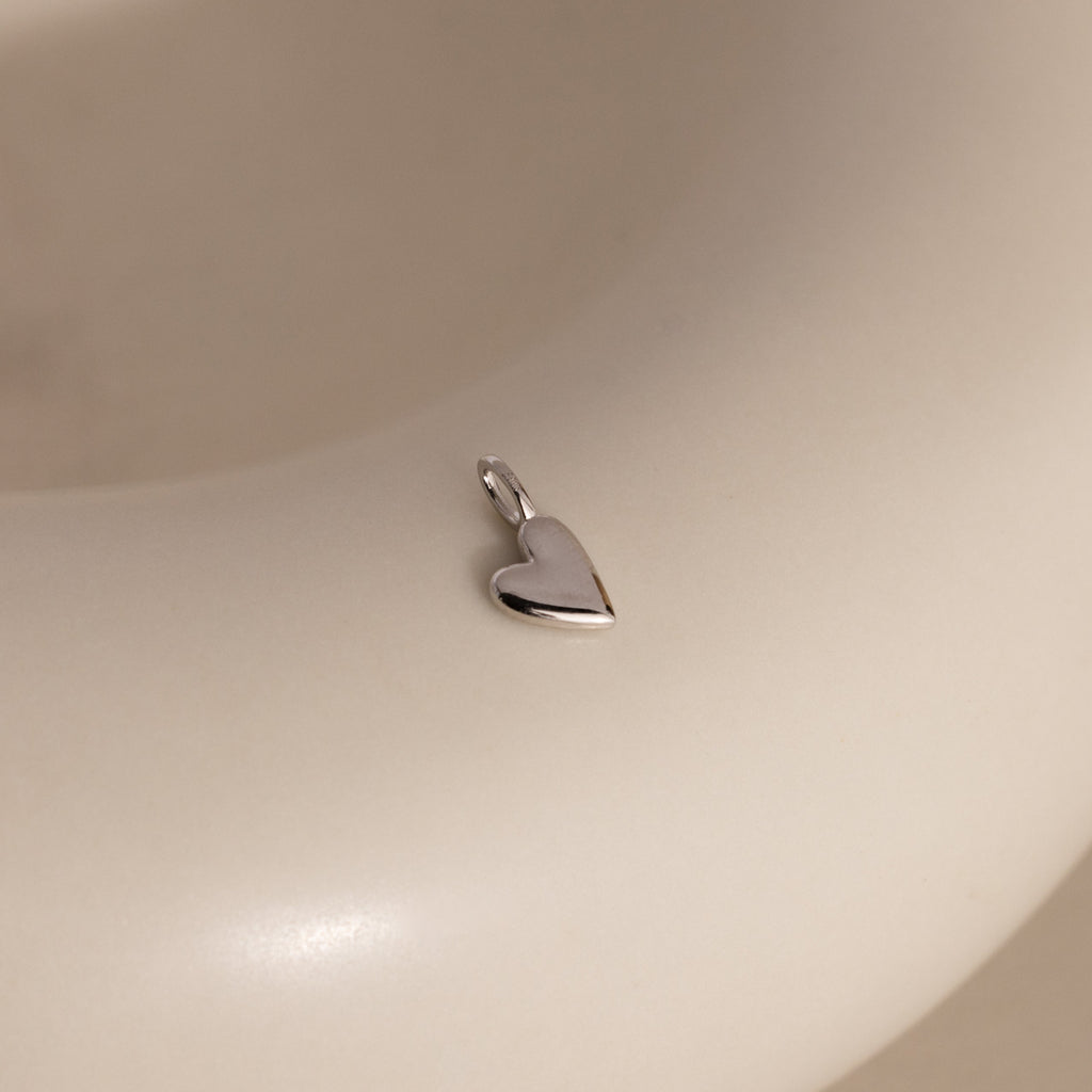 A silver Simple Heart Charm rests on a soft beige backdrop, its curved silhouette and high-polish finish catching the light.