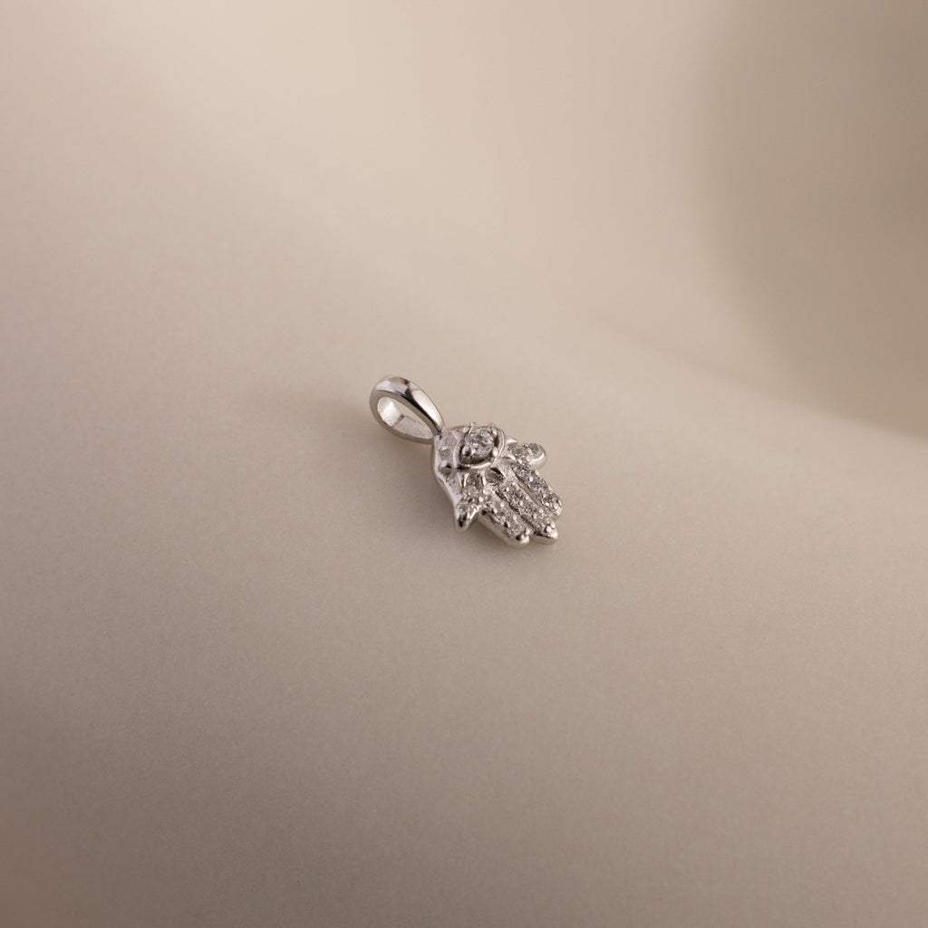 A close-up of the silver Evil Eye Hand Charm lying on a soft beige surface, showcasing its detailed stone-studded design.