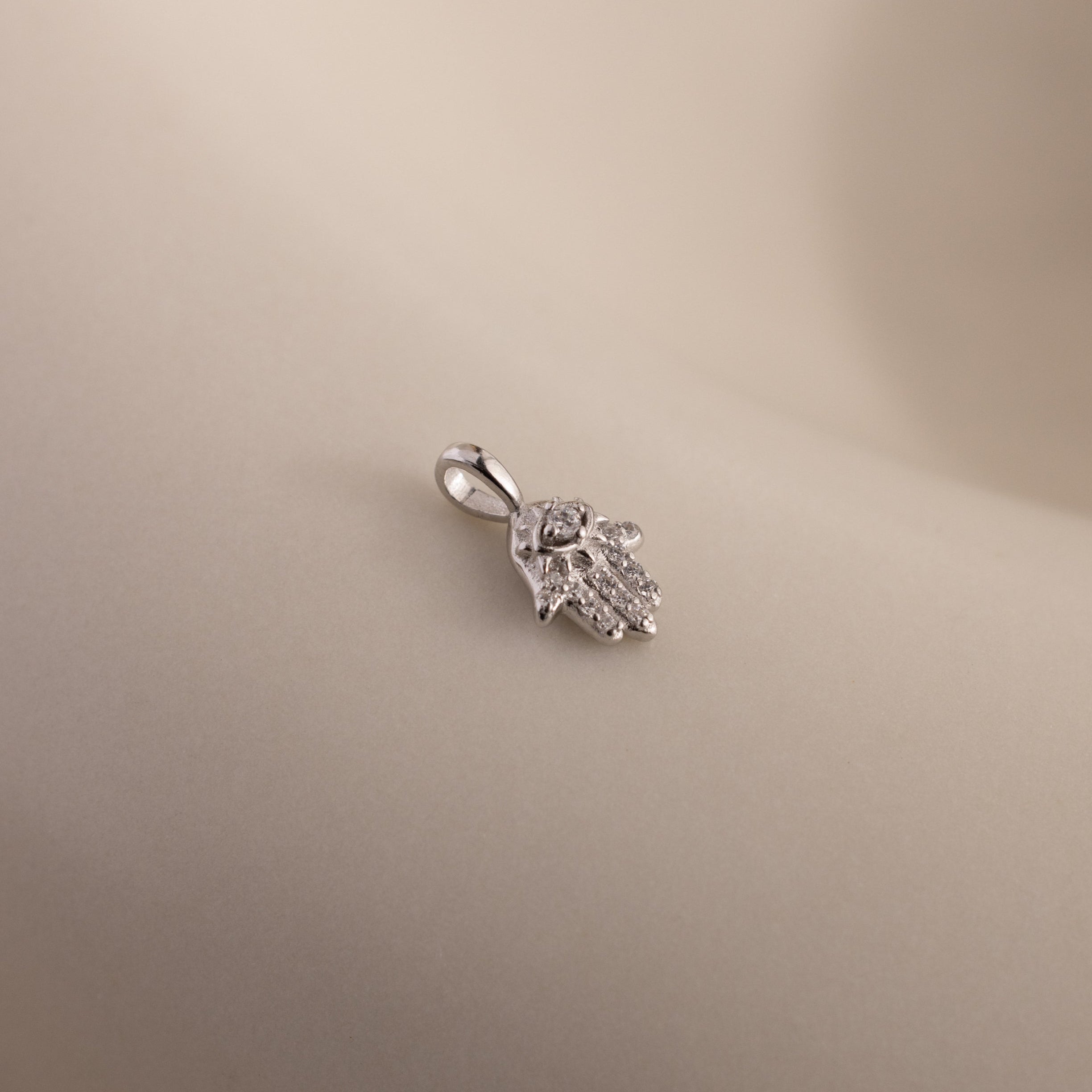 A close-up of the silver Evil Eye Hand Charm lying on a soft beige surface, showcasing its detailed stone-studded design.