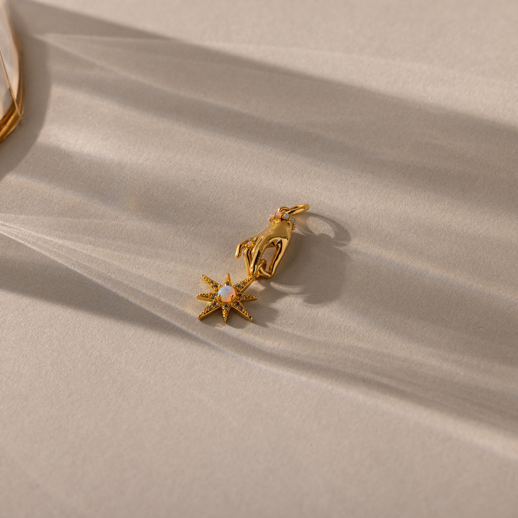 A close-up of a gold pendant featuring a sculpted hand holding an eight-pointed starburst charm set with an iridescent opal center