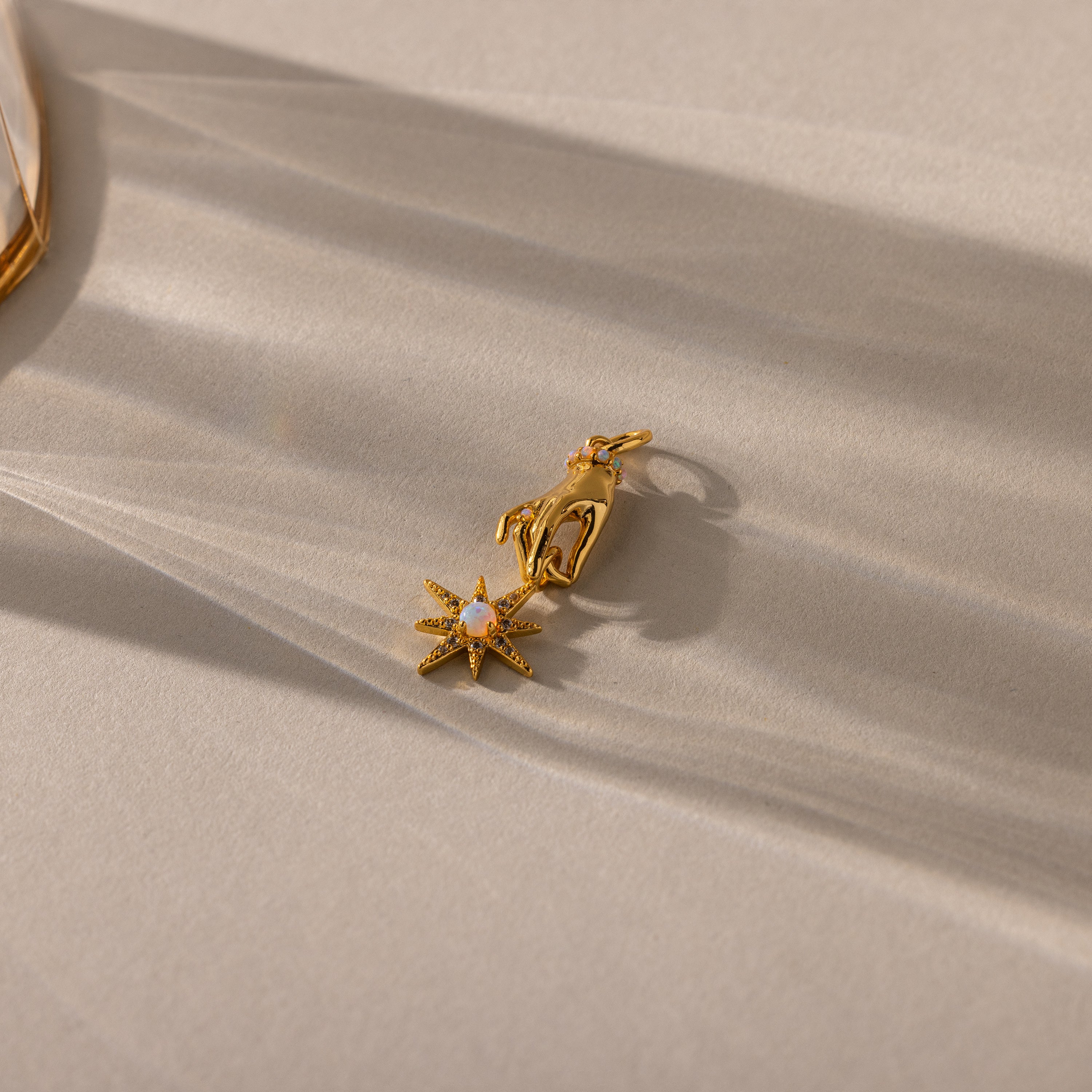A close-up of a gold pendant featuring a sculpted hand holding an eight-pointed starburst charm set with an iridescent opal center