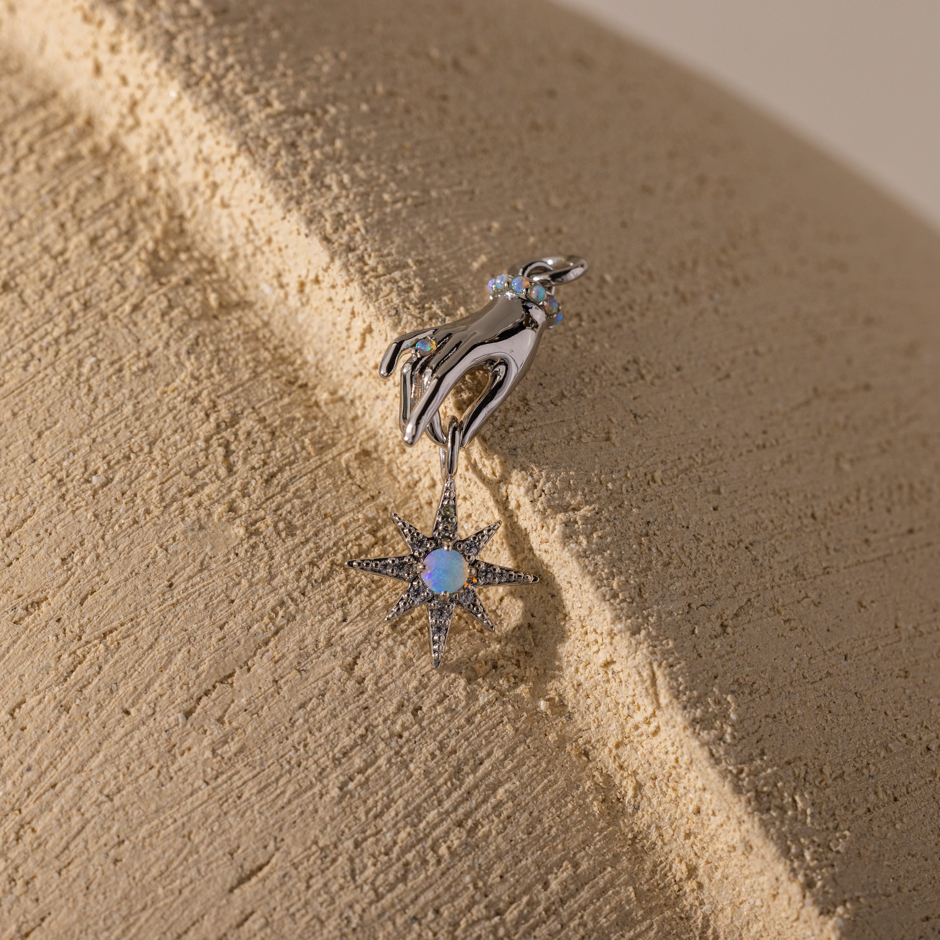 Silver charm of a hand holding an eight-pointed star with a glowing opal center, shown on a textured beige surface.
