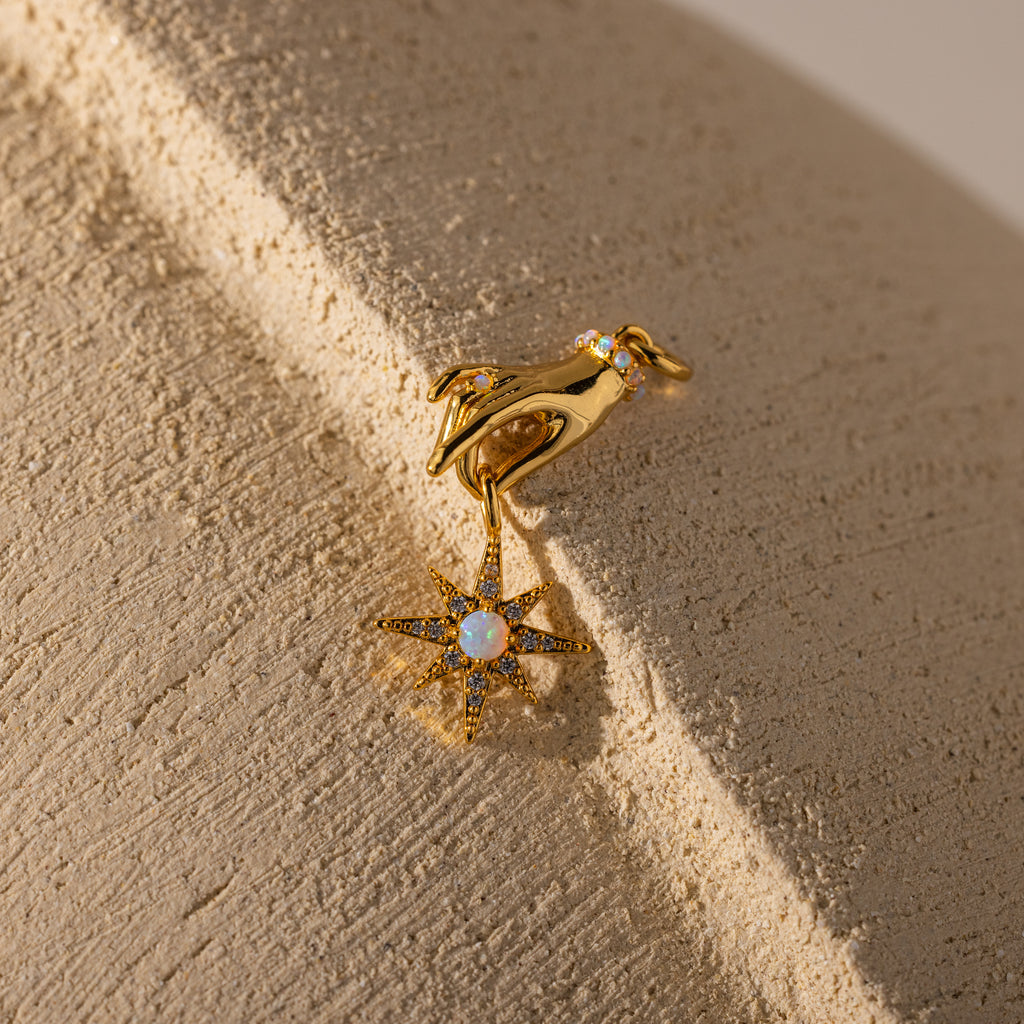 The Star Opal Hand Charm earring features a hand holding a star and opal stones, symbolizing intuition and cosmic energy, shown on a textured beige surface.