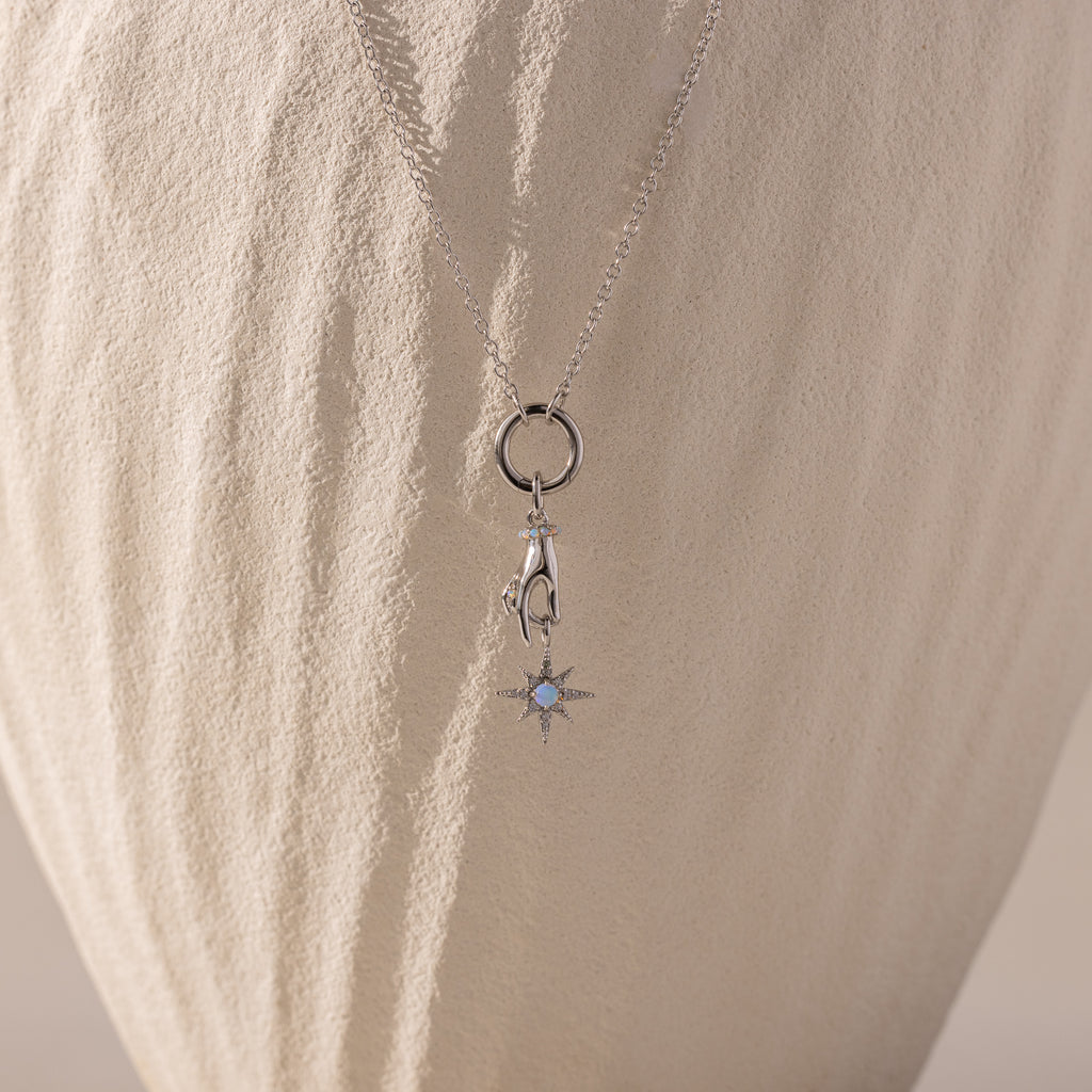 Silver necklace with a charm of a hand holding an opal-centered star, displayed against a textured, cream-colored background
