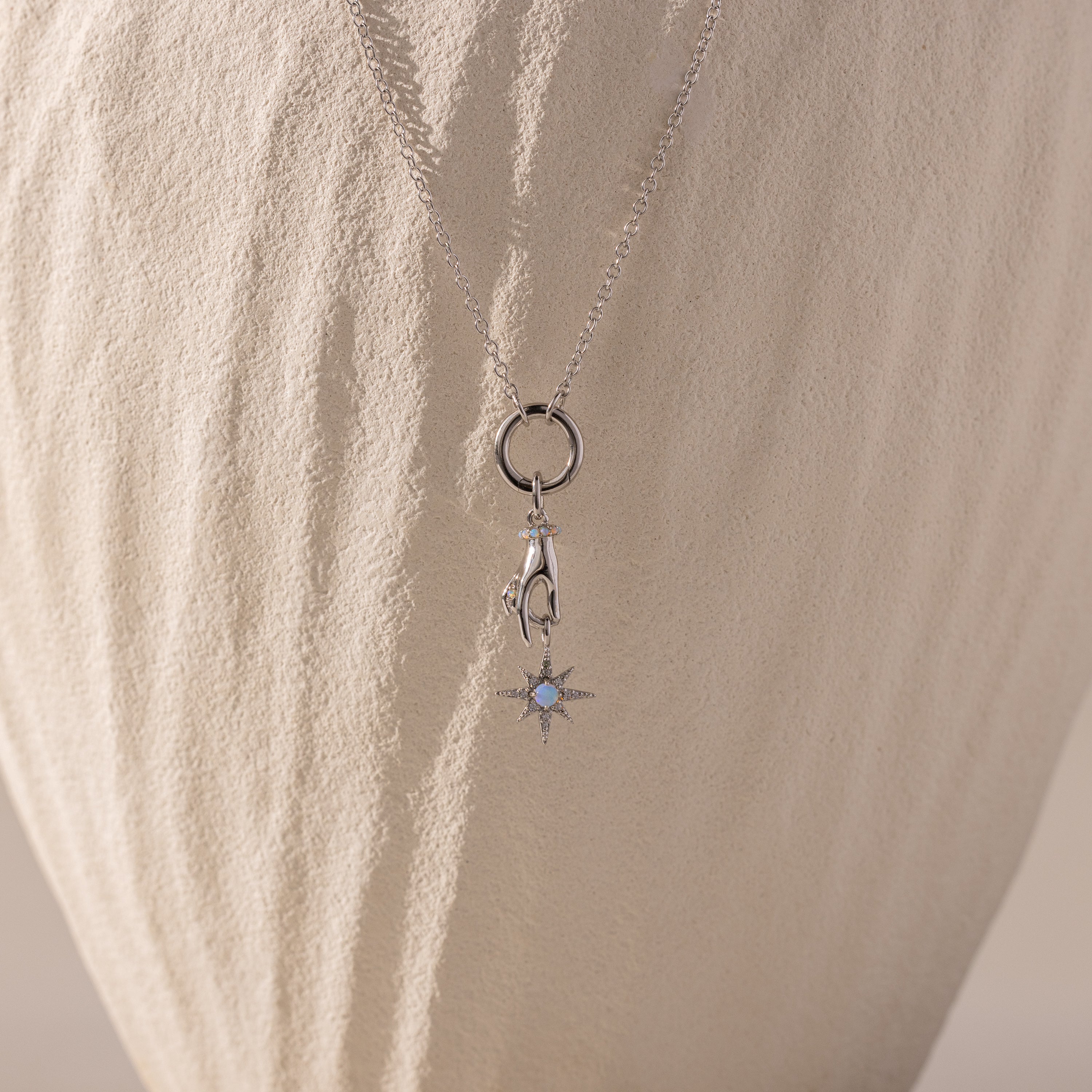 Silver necklace with a charm of a hand holding an opal-centered star, displayed against a textured, cream-colored background