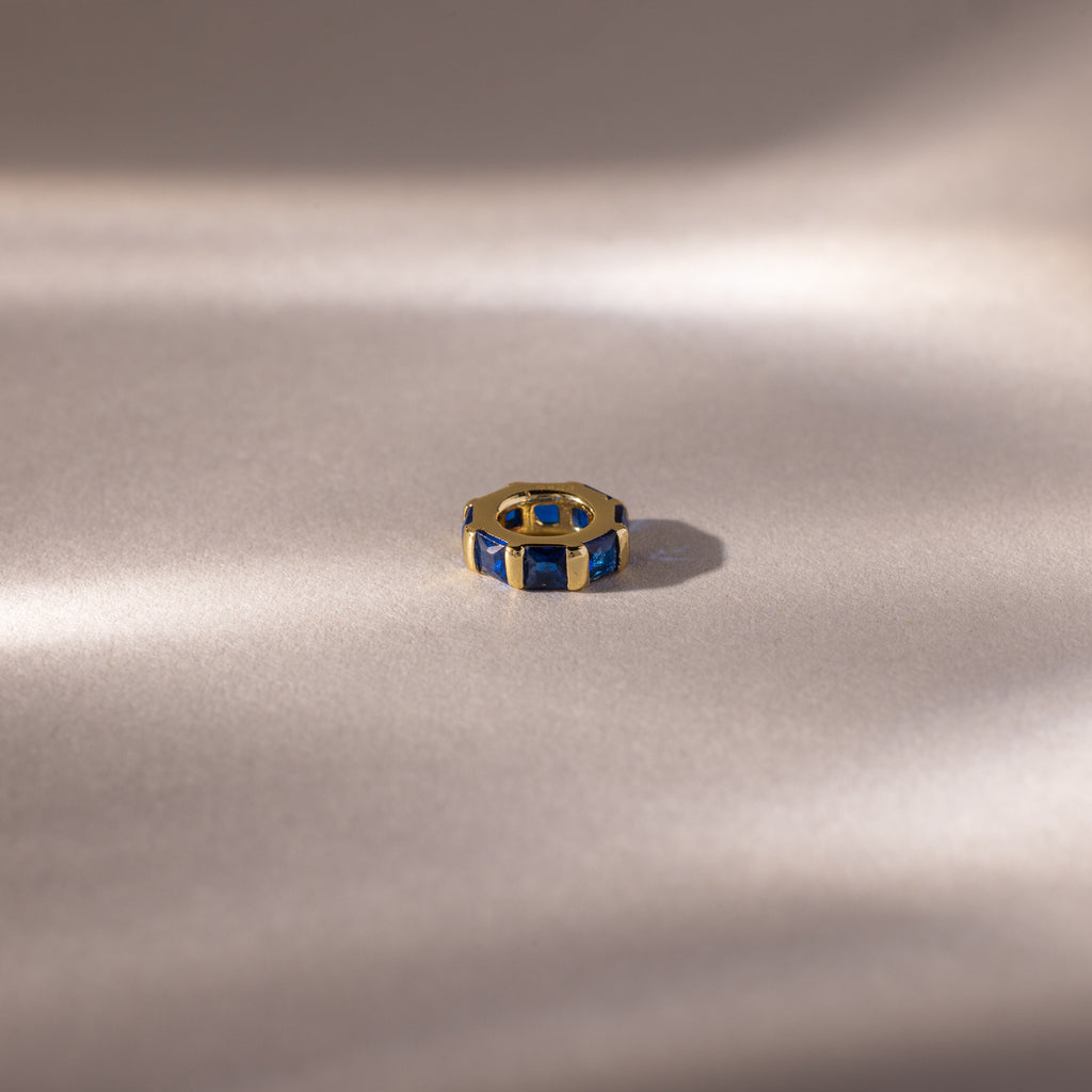 The Infinity Princess Sapphire Charm, a gold ring with princess-cut blue gemstones, is displayed on soft, light fabric, evoking elegance amid gentle shadows.