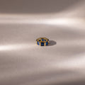 The Infinity Princess Sapphire Charm, a gold ring with princess-cut blue gemstones, is displayed on soft, light fabric, evoking elegance amid gentle shadows.