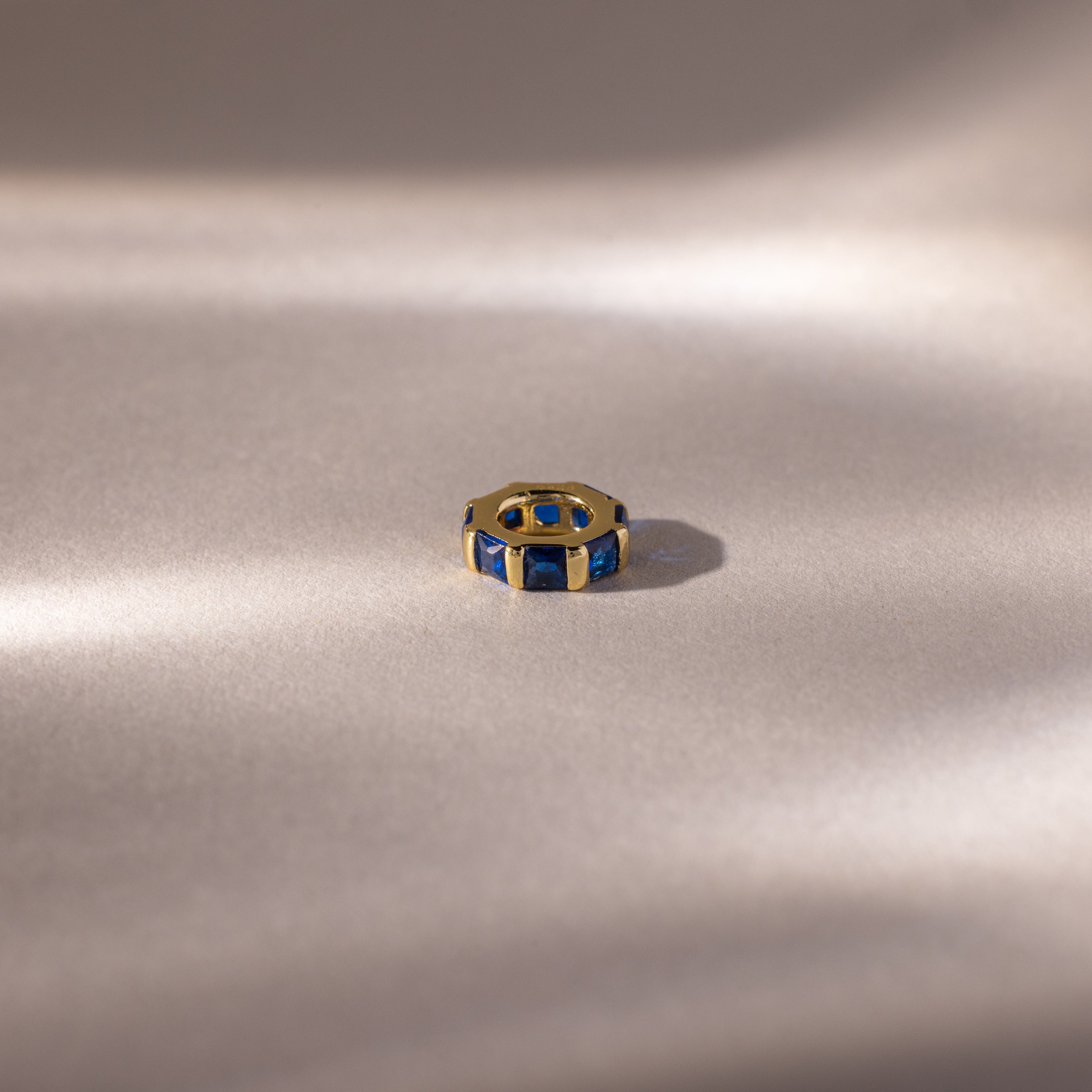 The Infinity Princess Sapphire Charm, a gold ring with princess-cut blue gemstones, is displayed on soft, light fabric, evoking elegance amid gentle shadows.