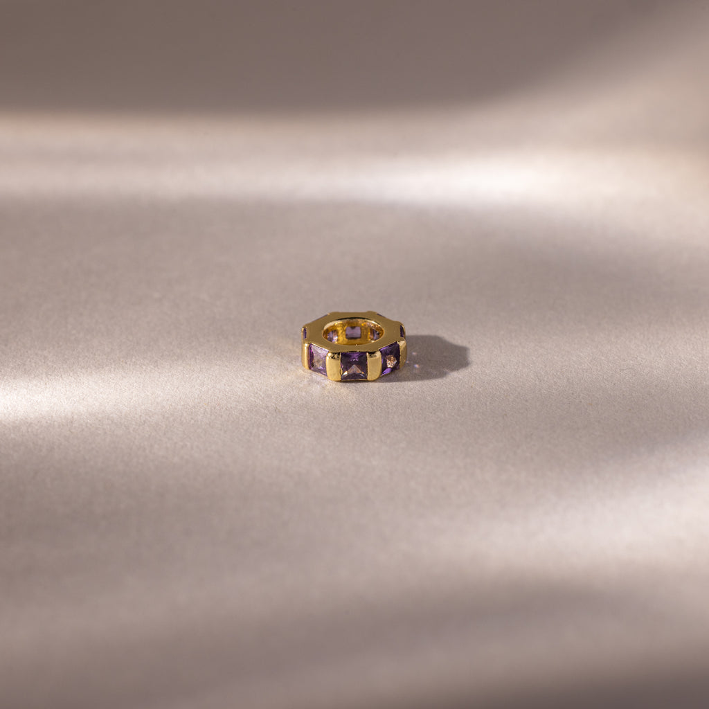 Gold ring with princess-cut, rich purple stones like the Infinity Princess Amethyst Charm, displayed on a soft, light backdrop with gentle shadows.