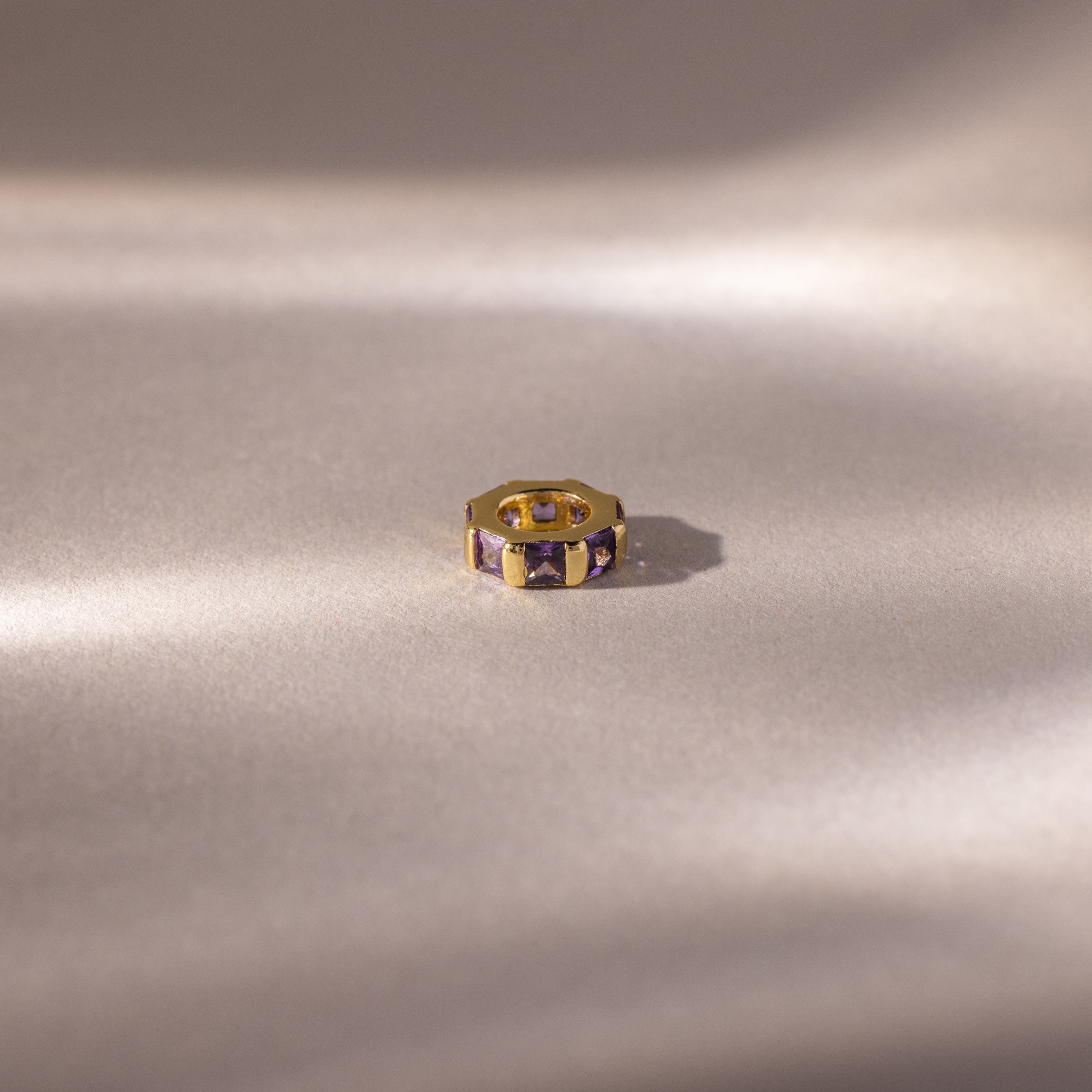 Gold ring with princess-cut, rich purple stones like the Infinity Princess Amethyst Charm, displayed on a soft, light backdrop with gentle shadows.