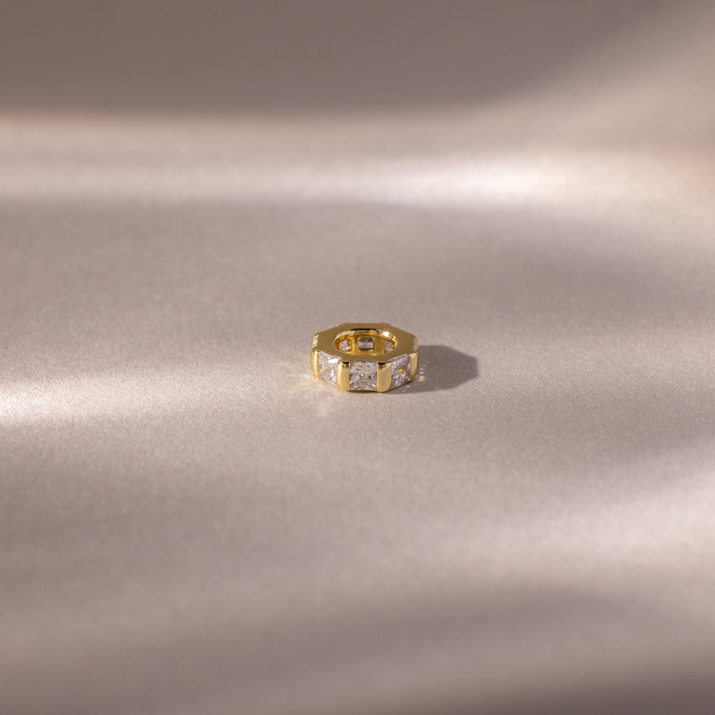 A gold ring with princess-cut stones, inspired by the Infinity Princess Diamond Charm, rests on a soft, light-colored satin surface.