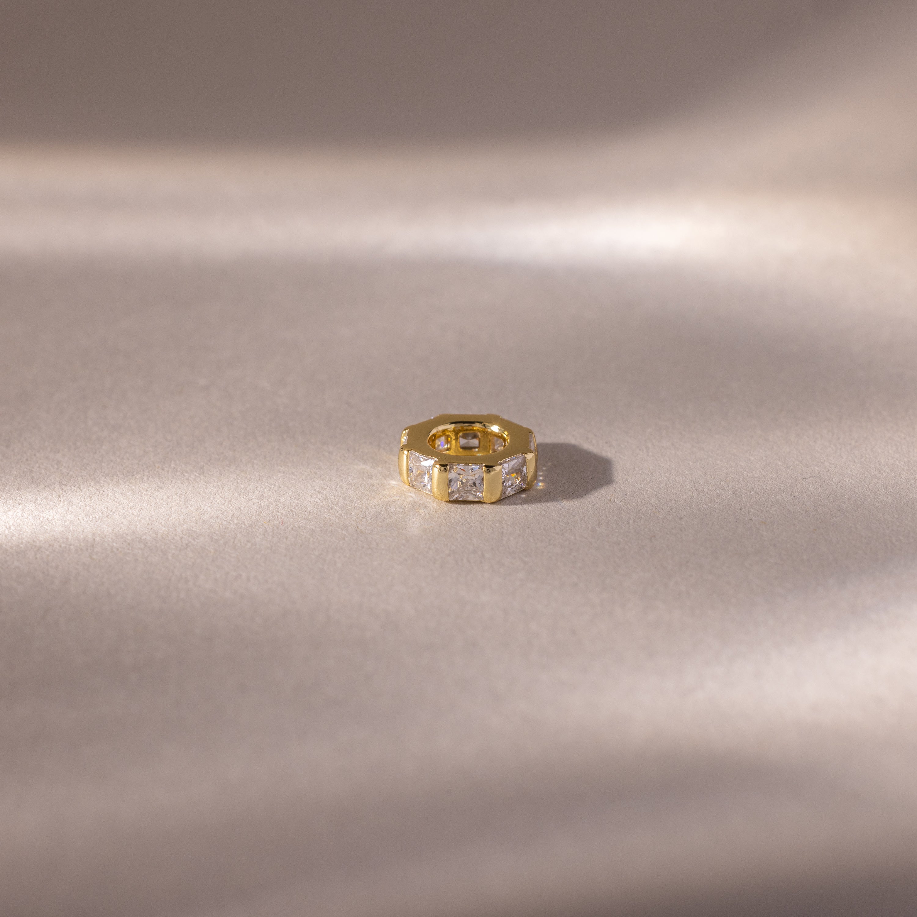 A gold ring with princess-cut stones, inspired by the Infinity Princess Diamond Charm, rests on a soft, light-colored satin surface.