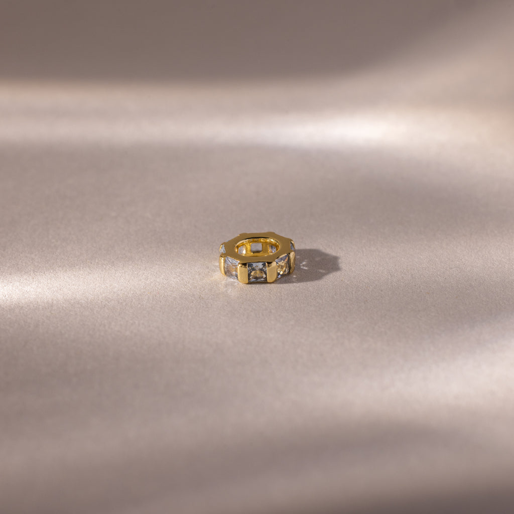 A gold ring with princess-cut stones rests on a soft beige surface with subtle shadows, evoking the elegance of the Infinity Princess Aquamarine Charm.