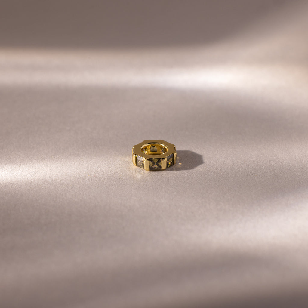 A small, gold hexagonal ring with an Infinity Princess Peridot Charm rests on a smooth, softly lit beige surface.